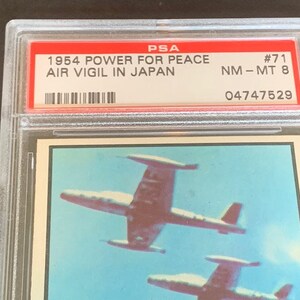 May include: A PSA graded card featuring a 1954 "Power for Peace" Air Vigil in Japan trading card. The card is graded NM-MT 8 and has the number 71. The card features a photo of two fighter jets flying in formation.