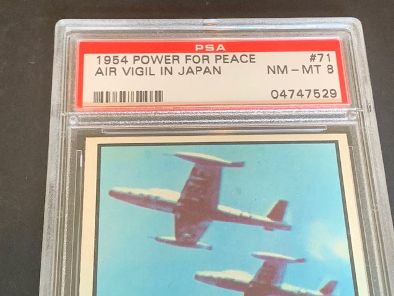 May include: A PSA graded card featuring a 1954 "Power for Peace" Air Vigil in Japan trading card. The card is graded NM-MT 8 and has the number 71. The card features a photo of two fighter jets flying in formation.
