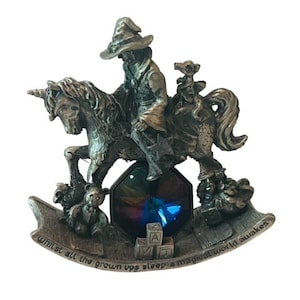 May include: A pewter figurine of a wizard riding a unicorn with a small dragon perched on the unicorn's back. The figurine is on a rocking horse base with a blue and iridescent crystal in the center. The base has the inscription "whilst all the grown ups sleep a magical world awakes".