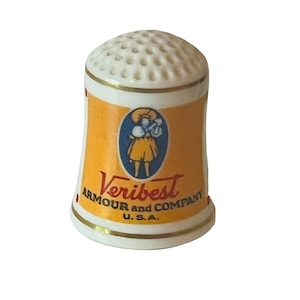 Op de afbeelding: Witte keramische vingerhoed met een geel, blauw en rood ontwerp. De vingerhoed heeft de tekst "Veribest Armour and Company U.S.A."