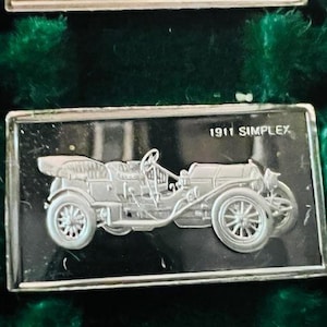 May include: A silver-toned rectangular bar with a black and white image of a 1911 Simplex car. The bar has a raised border and the text "1911 Simplex" is printed on the top.