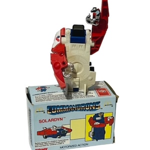 May include: A red, white, and blue toy robot with a motorized action feature. The robot is in its robot form and has a hook on its arm. The robot is in a box with the words "Commandrons" and "Solardyn" on it. The box also has a picture of the robot in its vehicle form.