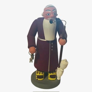 Könnte beinhalten: Eine Holzfigur eines Mannes mit weißem Bart und einem violetten Gewand. Er hält einen Stab und einen Schlüsselbund. Die Figur ist detailliert bemalt und hat einen gelben Sockel.