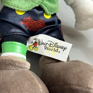 May include: Close-up of a plush toy featuring a green top, black pants with red floral designs, and brown shoes. The tag reads "Walt Disney World" with the Mickey Mouse logo.