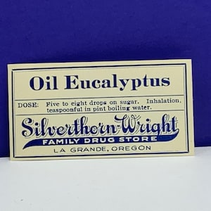 Pode incluir: Um rótulo vintage para Óleo de Eucalipto, um produto vendido pela Silverthorn-Wright Family Drug Store em La Grande, Oregon. O rótulo é impresso em papel creme com texto azul e uma borda azul.