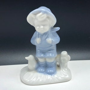 Puede incluir: Una figurita de porcelana de un niño con un sombrero y una chaqueta azules, de pie con un pájaro y un árbol detrás de él.