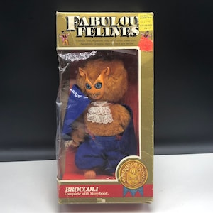 May include: A plush lion toy wearing a blue cape and a cat mask. The toy is packaged in a box with a gold background and the words "Fabulous Felines" printed on it. The box also includes the words "Cuddle 'em, squeeze 'em, don't ever tease 'em, Fabulous Felines, they're the Cats meow." and "BROCCOLI Complete with Storybook."
