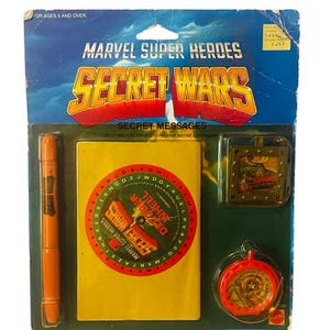 May include: A plastic package containing a Marvel Super Heroes Secret Wars Secret Messages set. The set includes a yellow notebook with a decoder wheel, a red and gold decoder wheel, a brown marker, and a gold metal keychain with a picture of a spaceship.