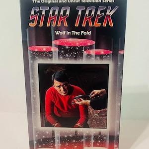 Puede incluir: Una caja de cinta VHS para el episodio de la serie de televisión Star Trek "Wolf in the Fold". La caja presenta un fondo rojo con una imagen en blanco y negro del Capitán Kirk con una camisa roja.