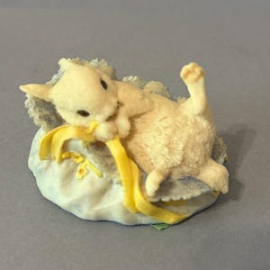 May include: A cream-colored figurine of a playful bunny rabbit. The bunny is lying on its back, playfully tugging at a yellow ribbon. The base is a light blue, cloud-like shape with a touch of green.