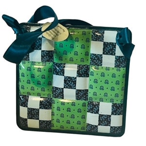 May include: A square, woven bag with a teal handle and bow. The bag features a checkerboard pattern of white, black, and green squares. The green squares have a small floral design. A small tag is attached to the bow.