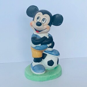 May include: A ceramic figurine of Mickey Mouse dressed in a blue and yellow soccer uniform, standing with his arms crossed and holding a black and white soccer ball.