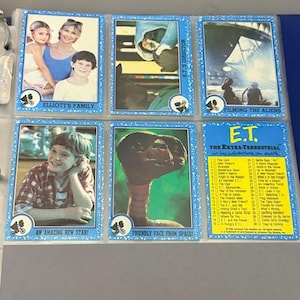 May include: A collection of six trading cards from the movie E.T. The Extra-Terrestrial, featuring images of the characters and scenes. The cards are in a blue border and include text such as "Elliott's Family" and "Friendly Face From Space!"