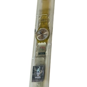 Puede incluir: Un reloj Swatch amarillo con una esfera blanca y una caja de plástico transparente. El reloj está en su embalaje original, que también es de plástico transparente. El embalaje tiene las palabras "Swatch Quartz" y "Swiss Made" impresas en él. El reloj tiene un logotipo olímpico de EE. UU. en la banda.