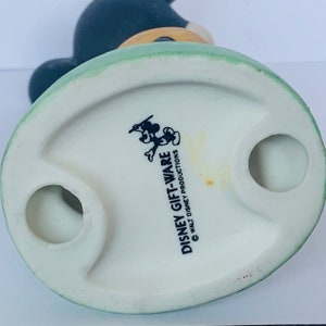 May include: White ceramic figurine of Mickey Mouse wearing a green hat and holding a paintbrush. The base of the figurine is oval shaped and has the text "DISNEY GIFT-WARE &copy; WALT DISNEY PRODUCTIONS" printed on it.