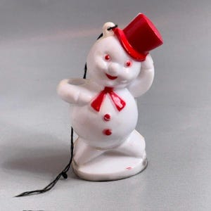 Könnte beinhalten: Eine weiße Schneemann-Figur mit glänzender Oberfläche, die einen leuchtend roten Zylinder und eine rote Fliege trägt. Der Schneemann hat rot bemalte Augen und zwei rote Knöpfe auf der Brust. Eine dünne schwarze Schnur ist am Hut befestigt.