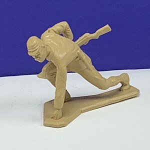 May include: A plastic toy soldier figurine in a crouched firing position, holding a rifle. The soldier is wearing a hat and has a detailed face. The figurine is tan in color and is standing on a base.