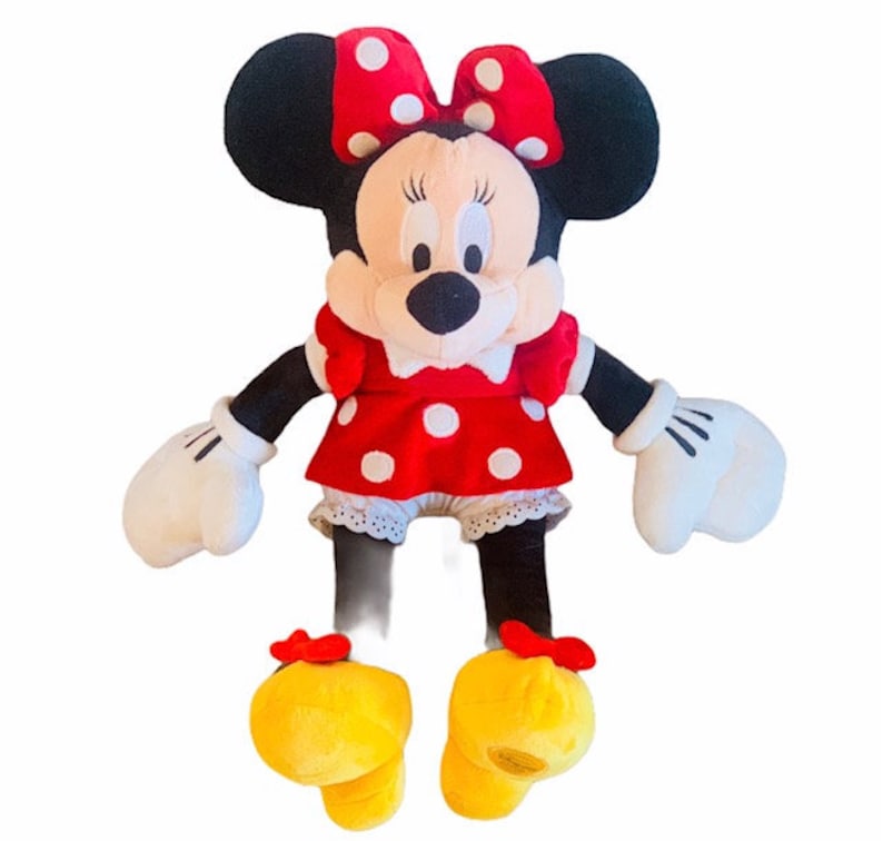 May include: A plush toy of Minnie Mouse wearing a red dress with white polka dots and a red bow with white polka dots. She has black ears and a black nose. Her shoes are yellow with red bows.