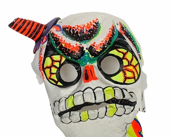 May include: A white paper mache skull mask with a bright green, yellow, orange, and black paint design. The mask has a large, toothy grin and a single horn on the top.