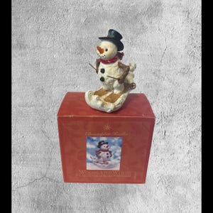 Könnte beinhalten: Eine weiße Schneemann-Figur, die einen schwarzen Hut und einen roten Schal trägt, auf einem schneebedeckten Hang Ski fährt. Die Figur ist Teil der Woodland Winds-Kollektion von Christopher Radko.