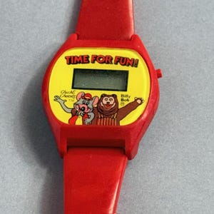 May include: A red digital wristwatch with a yellow face featuring the text "TIME FOR FUN!" and Chuck E. Cheese characters. The watch has a red strap and a digital display.