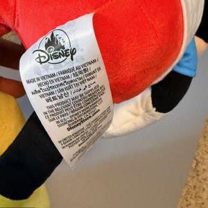 Pode incluir: Um brinquedo de pel&uacute;cia com as cores vermelho, preto, branco e azul. Uma etiqueta branca com o logotipo da Disney e texto em v&aacute;rios idiomas est&aacute; anexada. A etiqueta inclui o texto "MADE IN VIETNAM".