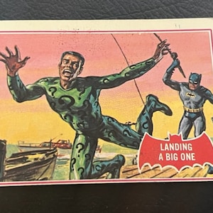 Könnte beinhalten: Eine Vintage-Batman-Sammelkarte mit dem Rätselraten in einem grünen Anzug mit einem Fragezeichen auf der Brust, der mit einer Angelrute in der Hand vom Himmel fällt. Batman ist im Hintergrund zu sehen, trägt einen blau-grauen Anzug und den Text "Landing a Big One" auf einem roten Banner.