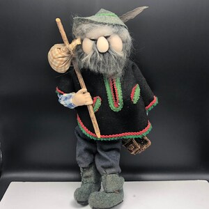 Puede incluir: Un muñeco de gnomo hecho a mano con una larga barba blanca, que lleva una túnica de fieltro negro y verde con ribetes rojos, un sombrero de fieltro verde con una pluma y sostiene un palo de madera. El gnomo lleva una pequeña bolsa a cuadros y una cesta.