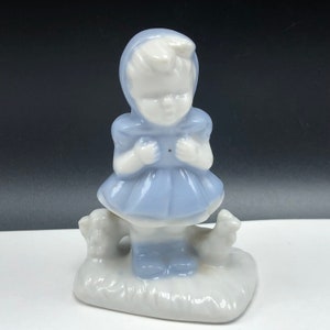 May include: A porcelain figurine of a girl in a blue dress and white bonnet standing with two small birds. The figurine is painted in shades of blue and white.