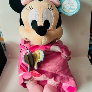May include: Pink Minnie Mouse plush toy wearing a pink and white blanket with a butterfly on the front. The toy has a pink bow on its head and is sitting on a white surface. The tag reads "Disney Babies".