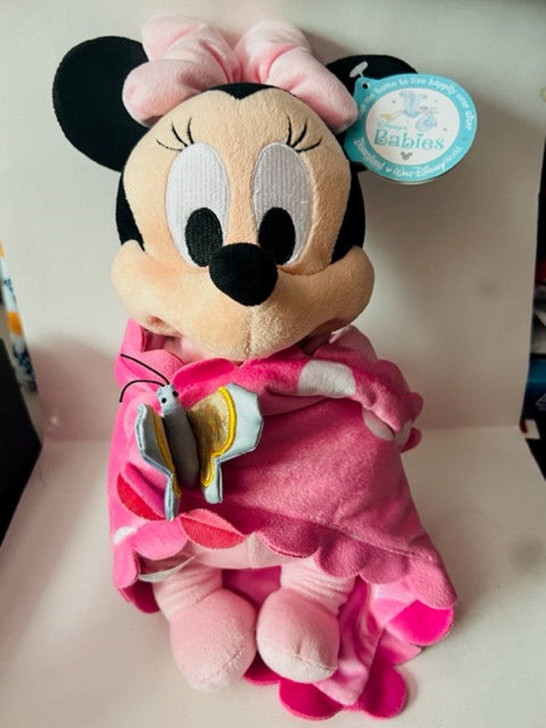 May include: Pink Minnie Mouse plush toy wearing a pink and white blanket with a butterfly on the front. The toy has a pink bow on its head and is sitting on a white surface. The tag reads "Disney Babies".