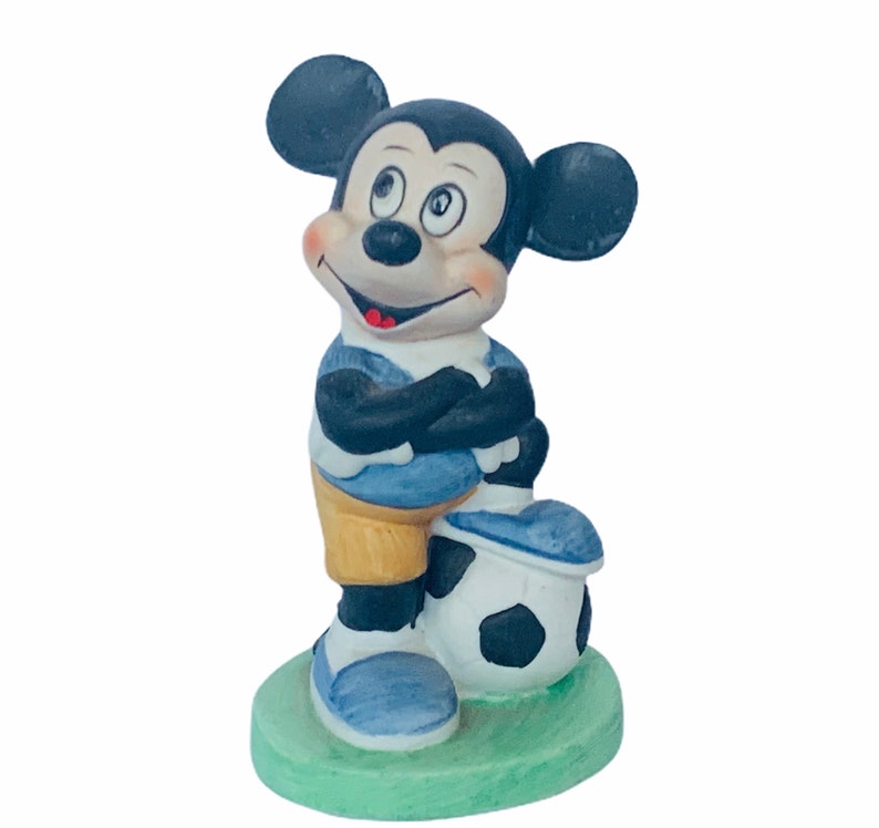 May include: A ceramic figurine of Mickey Mouse wearing a blue and yellow soccer uniform and holding a black and white soccer ball. The figurine is standing on a green base.