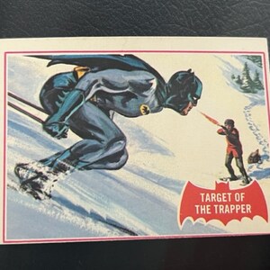 Könnte beinhalten: Eine Vintage-Sammelkarte mit einer Schwarz-Blauen Illustration von Batman, der einen schneebedeckten Berg hinunterskifährt. Die Karte hat unten ein rotes Banner mit der Aufschrift "Target of the Trapper".