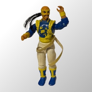 May include: A vintage action figure with a yellow and blue outfit, a black ponytail, and a yellow headband. The figure is holding a yellow rope in its right hand and has a yellow and blue wristband on its left arm.