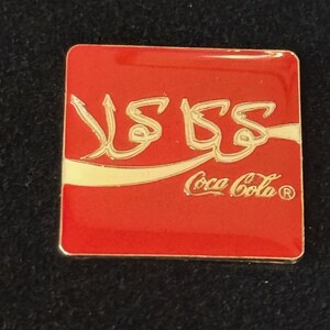 May include: A square, red Coca-Cola pin with gold lettering and a white curved stripe. The pin features the Coca-Cola logo in English and Arabic script. The pin is a collectible item.