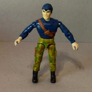 May include: A vintage action figure of a soldier in a blue sweater and camouflage pants. The figure has a brown strap across the chest and a blue knit cap. Black boots complete the outfit. The figure is posed with arms outstretched.