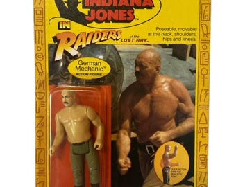 May include: Action figure packaging for the German Mechanic from the Indiana Jones Raiders of the Lost Ark movie. The packaging features a yellow background with a red border and the character's name in white text. The figure is shown in a plastic blister pack with a poseable action figure of the German Mechanic.