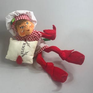 May include: A whimsical fabric doll in a chef's outfit, featuring a white chef's hat and a red and white checkered shirt. The doll holds a small pillow with the word "FLOUR" printed on it. The doll's legs and feet are bright red.