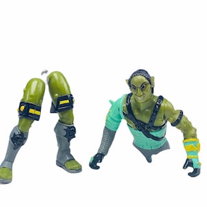 May include: Green and gray plastic action figure of a goblin with a green and blue torso, black straps, and yellow accents. The figure is missing its legs, which are shown separately.