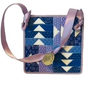 May include: A blue and purple patchwork quilt ornament with a lavender ribbon. The ornament has a gold tag that reads "The Quilting Dreams Collection, Phoenix, Grandvill".