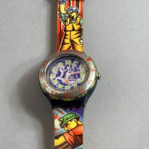 May include: Vintage wristwatch with a colorful design. The watch face features a blue and purple design with silver accents. The watch band is red, orange, and yellow with illustrations of a construction worker. The watch has a black case and a silver bezel.