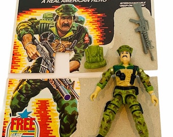 May include: A green and brown camouflage action figure of a soldier with a black gun and a green backpack. The figure is on a card with the words "GI JOE A REAL AMERICAN HERO" and "FREE INSIDE GI JOE ACTION FIGURE BODY TRANSFER".