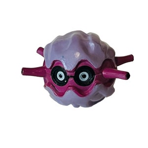 Op de afbeelding: Een paars en roze cartoonfiguur met grote ogen en twee kleine armen. Het figuur is gemaakt van plastic en heeft een textuur.
