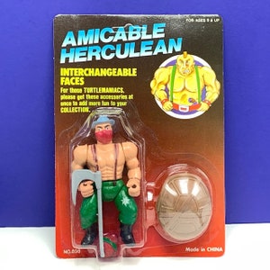 May include: An Amicable Herculean action figure on a red and black card. The figure is muscular, wearing green pants, a red mask, and a blue hat. It holds a gray axe and a red object. The card reads "INTERCHANGEABLE FACES."