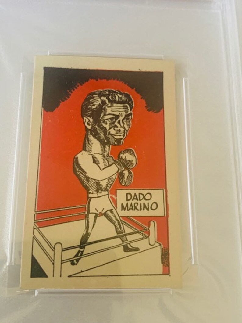 May include: A black and white cartoon illustration of a boxer in a boxing ring. The boxer is wearing boxing gloves and shorts. The text "DADO MARINO" is printed on a white rectangle in the upper right corner of the image.