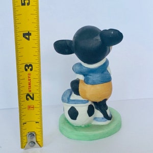 May include: A ceramic figurine of a cartoon character wearing a blue and yellow jersey, sitting on a soccer ball. The figurine is about 3 inches tall and is painted in black, white, blue, and yellow.