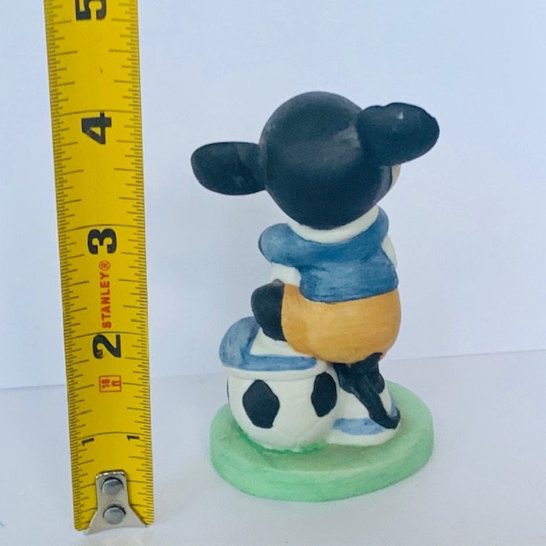 May include: A ceramic figurine of a cartoon character wearing a blue and yellow jersey, sitting on a soccer ball. The figurine is about 3 inches tall and is painted in black, white, blue, and yellow.