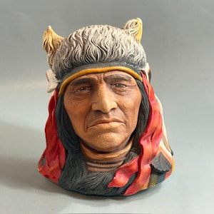 May include: A ceramic mug shaped like the head of a person wearing a textured gray fur headdress with small horns. The figure has dark hair, a tan complexion, and is adorned with bright red fabric accents and white feathers on the sides.