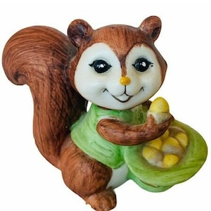 May include: A ceramic squirrel figurine with brown fur, green clothing, and a basket of yellow acorns. The squirrel has a smiling face and large, black eyes.