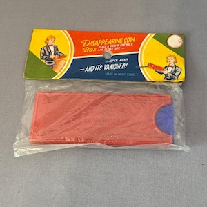May include: A red plastic box with a blue flap and the words "COIN CASE" printed on it. The box is packaged in a clear plastic bag with a colorful label that reads "DISAPPEARING COIN BOX" and "OPEN AGAIN - AND ITS VANISHED!" Made in Hong Kong.